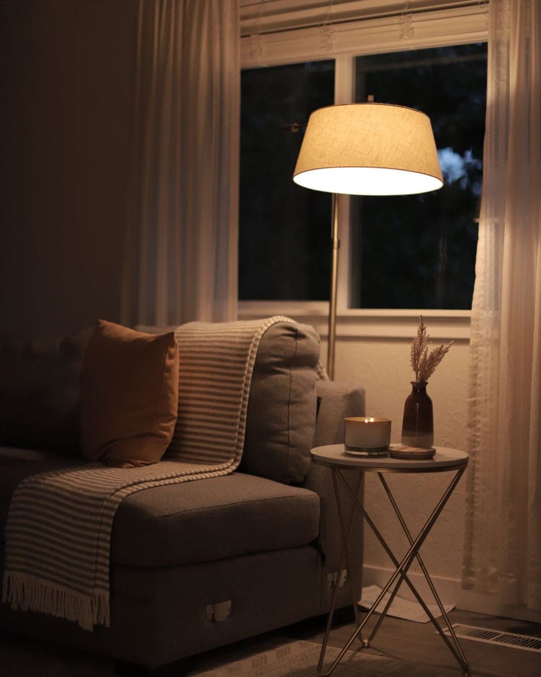 A quiet living-room corner at night: a floor lamp glowing over a sofa with a throw, and a candle on a side table